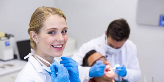 Zahnärztin mit blauen Handschuhen lächelt in die Kamera, während im Hintergrund ein Zahnarzt einen Patienten behandelt.