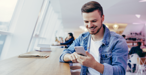 Junger Mann sitzt lächelnd an einem Tisch und bedient sein Smartphone.