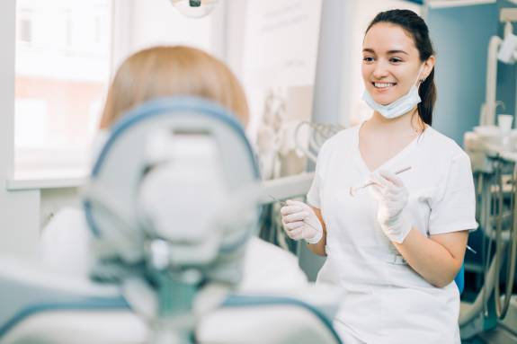 Zahnärztin mit Handschuhen und Mundschutz führt ein Beratungsgespräch mit einer Patientin im Behandlungsstuhl.