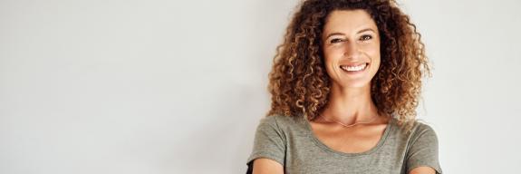 Porträt einer lächelnden Frau mit lockigem Haar und verschränkten Armen vor hellem Hintergrund Porträt einer lächelnden Frau mit lockigem Haar und verschränkten Armen vor hellem Hintergrund