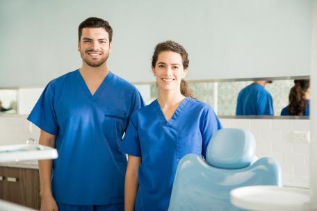 Lächelndes Zahnarztteam in blauer Praxiskleidung neben einem Behandlungsstuhl in einer modernen Zahnarztpraxis Lächelndes Zahnarztteam in blauer Praxiskleidung neben einem Behandlungsstuhl in einer modernen Zahnarztpraxis